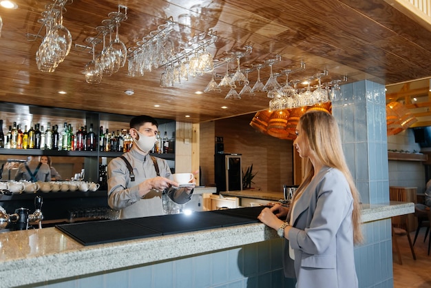 Een gemaskerde barista bereidt en serveert heerlijke natuurlijke koffie aan een jong meisje in een prachtig café tijdens een pandemie