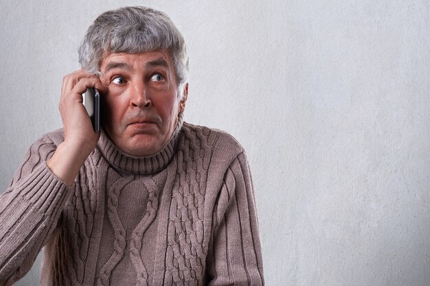 Een gepensioneerde die telefonisch spreekt en verbaasd is om een telefoontje te ontvangen. Een verbaasde uitdrukking van volwassen man in trui geïsoleerd over witte muur met mobiele telefoon in zijn hand.