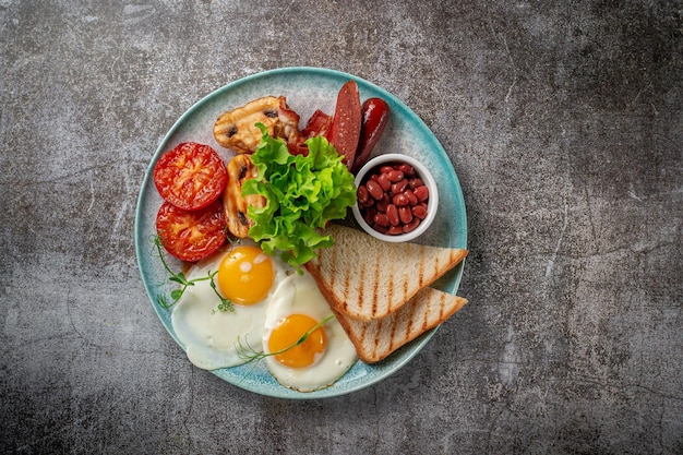 Een gerecht serveren van de menukaart van het restaurant. Heerlijk en gezond ontbijt met gebakken eieren, bonen, worstjes, gegrilde groenten, tomaten en champignons met salade op een bord tegen een grijze stenen tafel