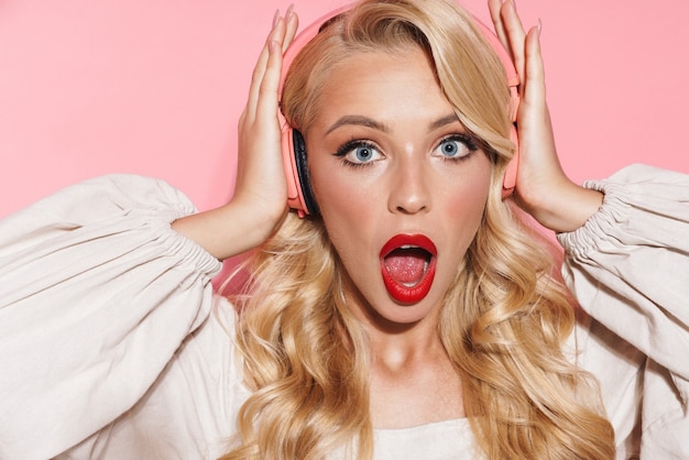 een geschokte jonge blonde vrouw die muziek luistert met een koptelefoon poseren geïsoleerd over roze muur muur.