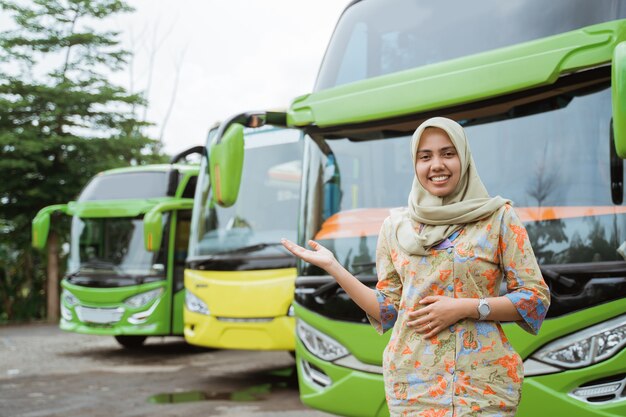 Een gesluierde vrouwelijke busbemanning die met handgebaren glimlacht en iets aanbiedt tegen de achtergrond van de busvloot