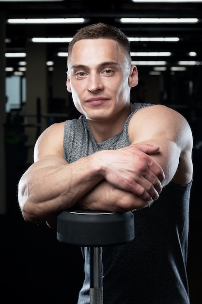 Een gespierde man in een grijs mouwloos shirt poseren leunend op een staande halter in een sportschool