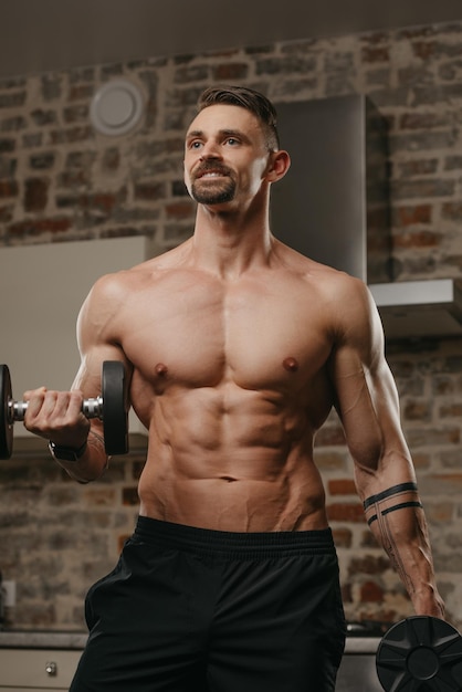 Foto een gespierde man met een baard traint zijn biceps met halters in zijn appartement een gelukkige bodybuilder met tatoeages op onderarmen doet thuis een armtraining een atleet met een naakte torso