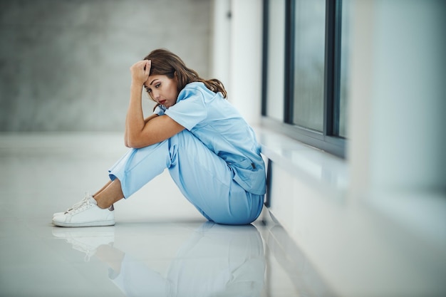 Een gestresste jonge verpleegster zittend op de vloer bij een raam terwijl ze een korte pauze heeft in een lege ziekenhuisgang tijdens de Covid-19-pandemie.