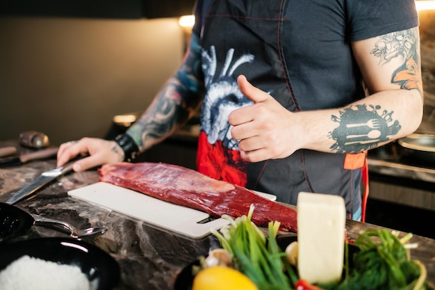 Een getatoeëerde chef-kok die een duim omhoog steekt voordat hij thuis sappige steak begint te koken Rundvlees op een snijplank