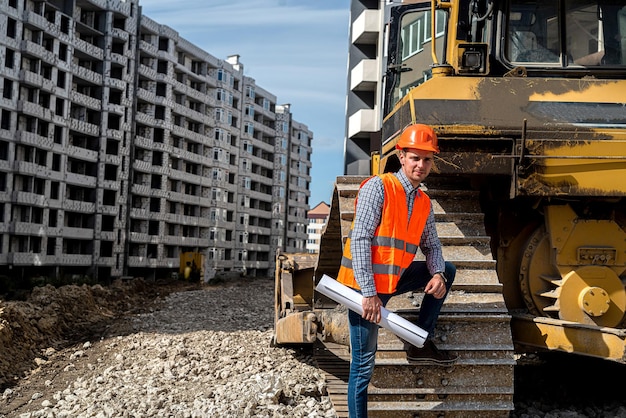 Een geweldige werker in uniform en helm staat bij een graafmachine op een bouwplaats Een graafmachine is in aanbouw Bouwconcept