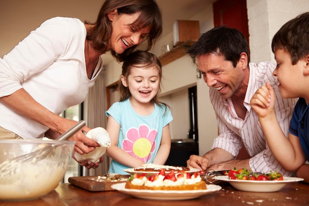 Een gezin met twee ouders en twee kinderen in de keuken die een taart met fruit en room glazuur maakt