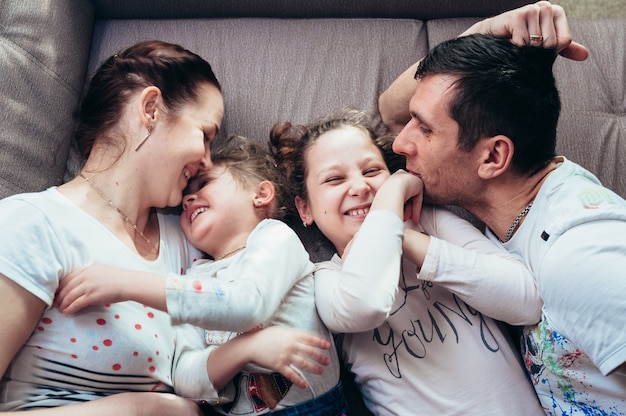 Een gezin van vier met gekrulde kinderen knuffelt en lacht terwijl ze op een grijze bank ligt