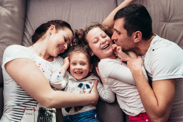 Een gezin van vier met gekrulde kinderen knuffelt en lacht terwijl ze op een grijze bank ligt