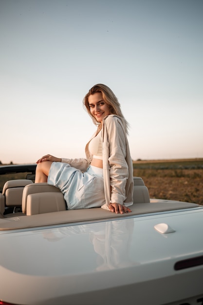 Een glamoureus luxe meisje met blond haar dat lacht terwijl ze op een witte cabrio zit. jonge succesvolle vrouw zit in haar witte auto