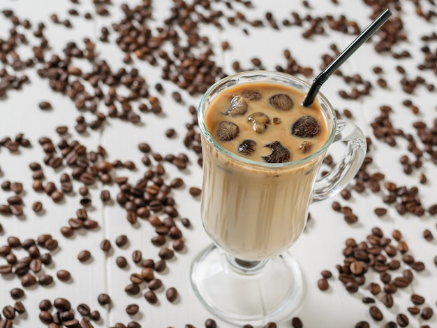 Een glas ijskoffie en een cocktail buis op de achtergrond van verspreide koffiebonen op een witte tafel. Verfrissende en verkwikkende drank van koffiebonen en melk.