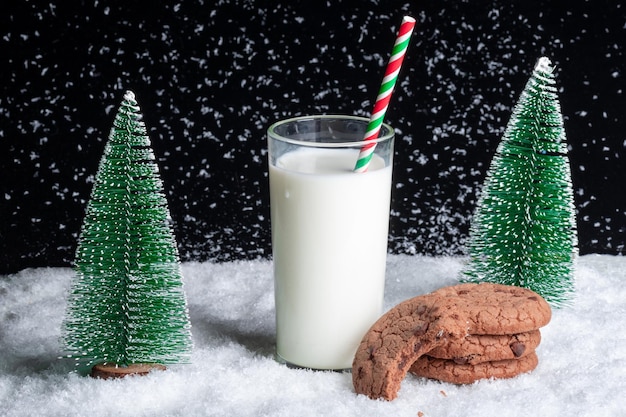 Een glas melk met een rietje en een half opgegeten koekje op een besneeuwde nachtelijke achtergrond tussen speelgoedkerstbomen