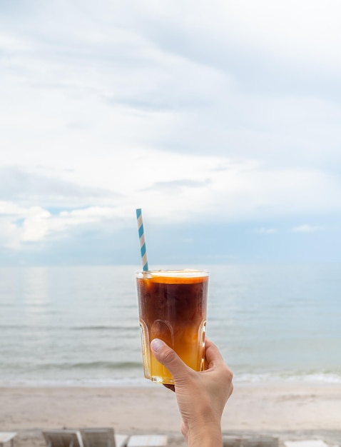 Een glas verse iced orange americano versierd met gesneden sinaasappel met de hand vasthouden op het strand uitzicht zeegezicht verticale stijl Koude zwarte koffie met sinaasappelsap mixen mocktail drankmenu