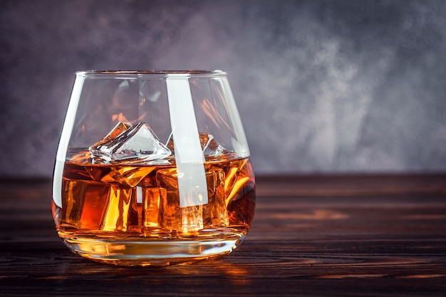 Een glas whisky op de donkerbruine houten tafel. Transparante gele alcoholische drank met ijs. Brandewijn, bourbon. Sterke alcoholische drank. Kopieer ruimte voor tekst, sjabloon.