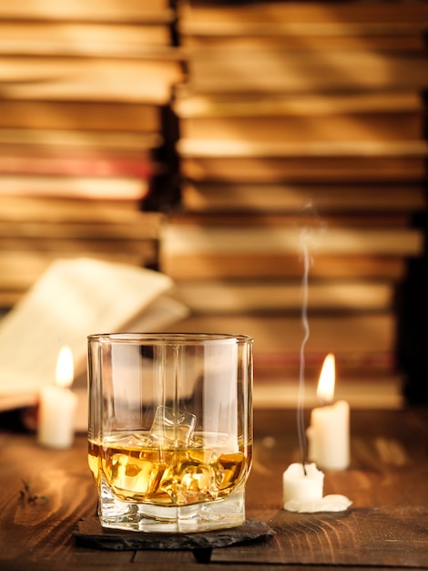 Een glas whisky op tafel met boeken en brandende kaarsen en lichtstralen. Kopieer ruimte, verticale foto.