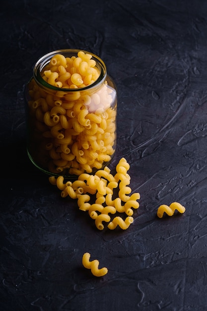Een glazen pot met cavatappi ongekookte gouden tarwe gekrulde pasta in de buurt van verspreide macaroni op getextureerde donkere zwarte muur, hoekweergave