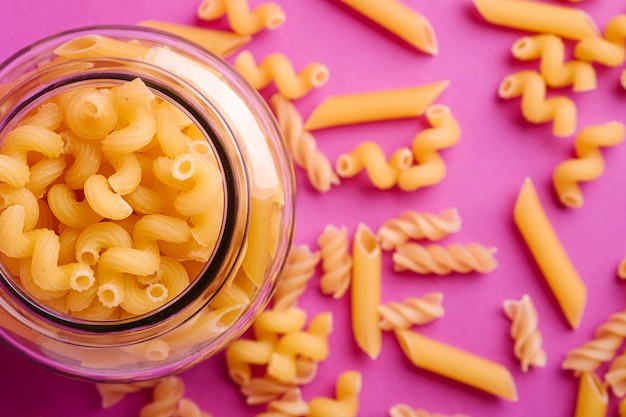 Een glazen pot met een verscheidenheid aan ongekookt gouden tarwe pasta op minimale roze achtergrond, bovenaanzicht macro