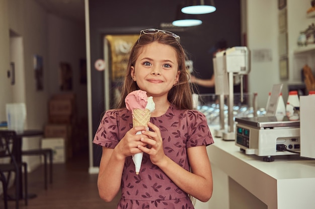 Een glimlachend schoonheidsmeisje in een modieuze jurk houdt aardbeienijs vast en staat in een ijssalon.