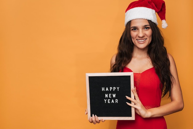 Een glimlachende blanke vrouw in een rode kerstmuts met een letterbord met het opschrift Happy New Year erop Op een gele geïsoleerde achtergrond Een plek voor uw tekst