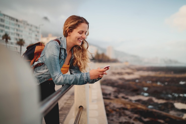 Foto een glimlachende blonde jonge vrouwelijke reiziger met haar rugzak die leunt op de reling op een stadsstraat met haar mobiele telefoon