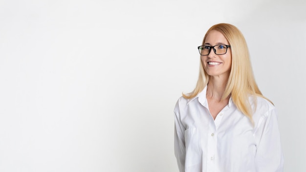Een glimlachende blonde vrouw met een zwarte bril en een wit hemd staat tegen een wit