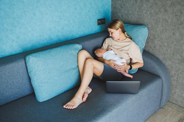 Een glimlachende jonge moeder geeft borstvoeding aan haar baby en werkt thuis met haar baby op de bank terwijl ze een e-mail schrijft Borstvoeding aan de baby Een jonge vrouw werkt thuis