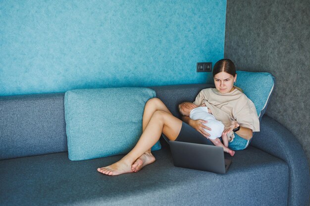 Een glimlachende jonge moeder geeft borstvoeding aan haar baby en werkt thuis met haar baby op de bank terwijl ze een e-mail schrijft Borstvoeding aan de baby Een jonge vrouw werkt thuis