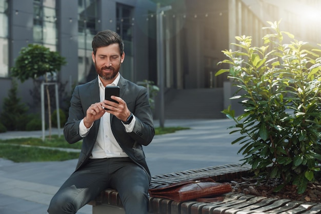 Een glimlachende kantoormedewerker rust tijdens de pauze buiten kantoor en scrollt door sociale media op de telefoon