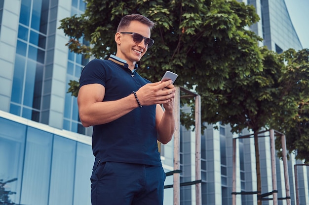 Een glimlachende knappe modieuze man met een stijlvol kapsel in zonnebril, gekleed in een zwart t-shirt en broek, leest een bericht op de smartphone, staat in de moderne stad tegen een wolkenkrabber.