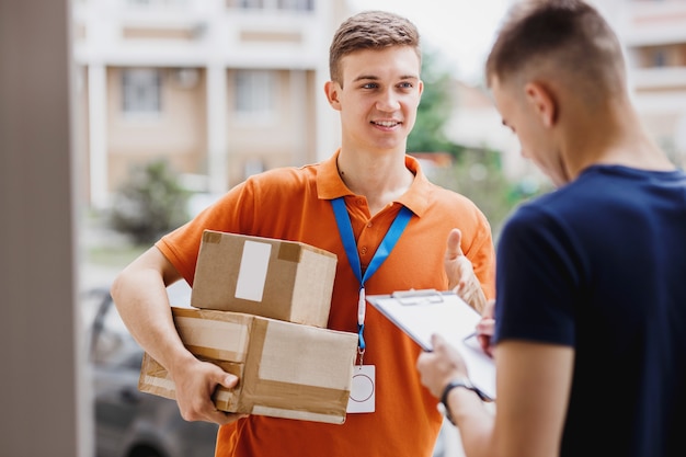 Een glimlachende persoon met een oranje T-shirt en een naamplaatje bezorgt een pakket aan een klant, die zijn handtekening op de bon zet. Vriendelijke werker, bezorgservice van hoge kwaliteit.