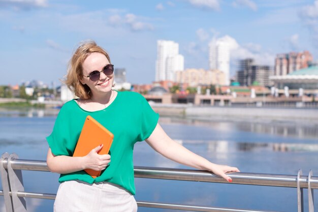 Een glimlachende vrouw van middelbare leeftijd in een zonnebril met een notitieboekje of tablet in haar handen op de stadsdijk