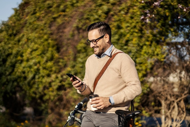 Een glimlachende zakenman leunt op zijn fiets in het park en drinkt afhaalkoffie en gebruikt zijn telefoon