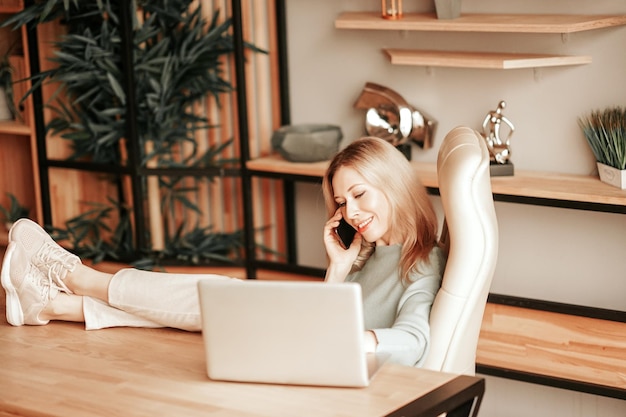 Een glimlachende zakenvrouw die op de stoel in haar kantoor zit en in de laptop kijkt en aan de telefoon praat