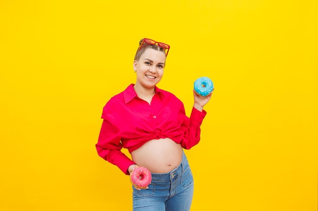 Een glimlachende zwangere vrouw in een roze shirt houdt donuts in haar handen op een gele achtergrond Zoet voedsel tijdens de zwangerschap Schadelijk voedsel tijdens de zwangerschap