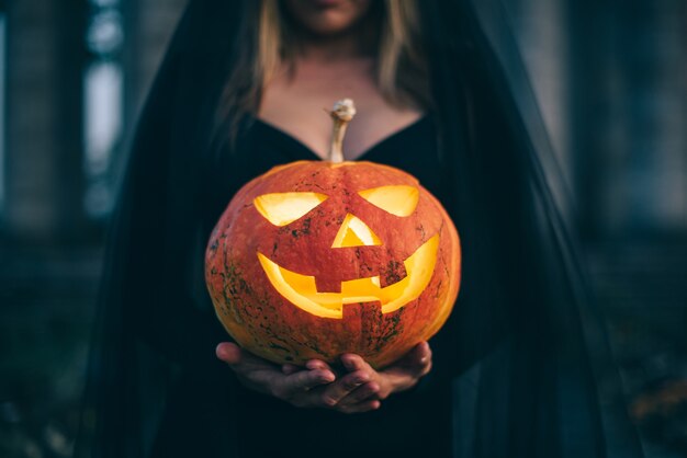 Een gloeiende pompoen met een vreselijk gezicht in de handen van een heks, close-up. Halloween-concept.