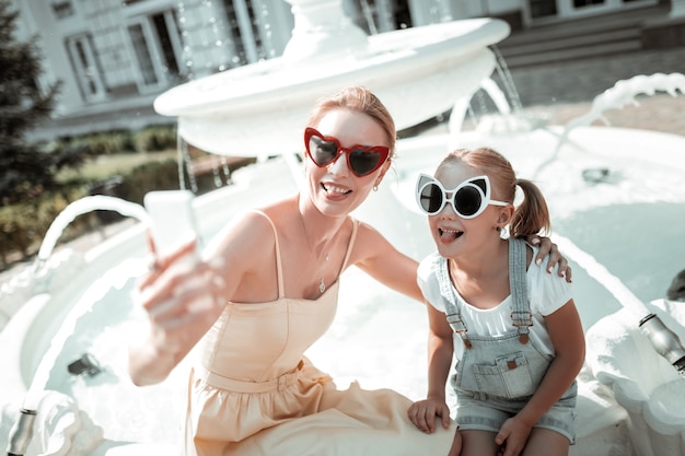 Een goede tijd hebben. Vrolijke moeder en dochter zitten in de buurt van de prachtige fontein en maken grappige gezichten die selfie nemen.