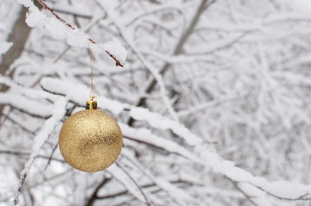 Een gouden speelgoedballon hangt aan een tak in de sneeuw op een wazige witte achtergrond met een plek voor tekst