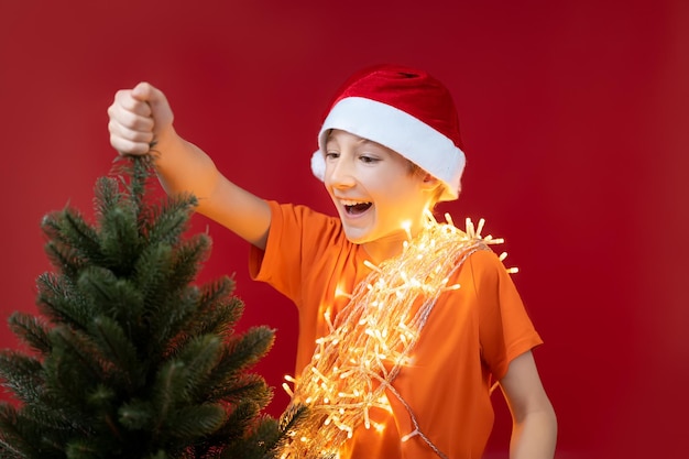 Een grappige jongen met een kerstmuts houdt de bovenkant van de kerstboom vast en lacht vrolijk