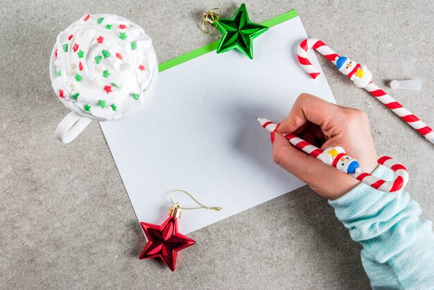 Een grijze tafel met een begroetingsvel, kerstversiering, een kop warme chocolademelk en een pen in de vorm van snoepgoed. Het schrijven van het meisje, wijfje dient beeld, hoogste mening in