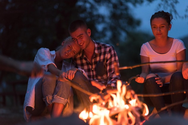 een groep gelukkige jonge vrienden die ontspannen en genieten van een zomeravond rond het kampvuur aan de oever van de rivier