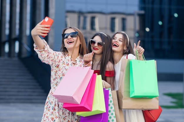 Een groep jonge mooie meisjes in vrijetijdskleding met zonnebril, make-up, haar hoepel en gekleurde boodschappentassen selfie maken na succesvol winkelen