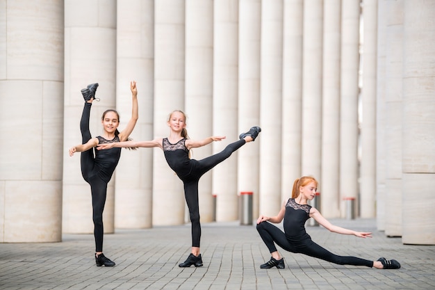 Een groep van drie kleine ballerina's in zwarte nauwsluitende kostuums danst tegen een achtergrond van het stadsbeeld.