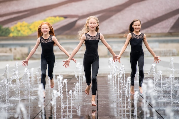 Een groep van drie kleine ballerina's in zwarte strakke pakken rennen naar de kijker te midden van spattende fonteinen tegen de achtergrond van een stadsbeeld op een warme dag.