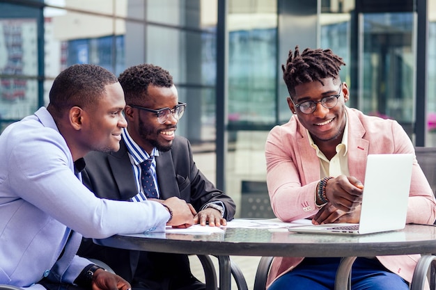 Een groep van drie stijlvolle Afro-Amerikaanse studenten-ondernemers in modepakken die aan tafel werken met een laptop in een zomercafé buitenshuis