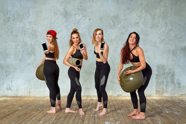 Een groep van mooie fitness meisjes in een fitnessruimte in de buurt van een grijze muur met een rekwisieten voor training, emotioneel praten aan de telefoon. Kopieer ruimte, grijze muur. Concept sport, smartphones