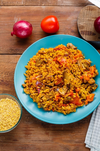 Een groot gerecht van bulgur met groenten en champignons in kruiden op een blauw servet naast tomaten