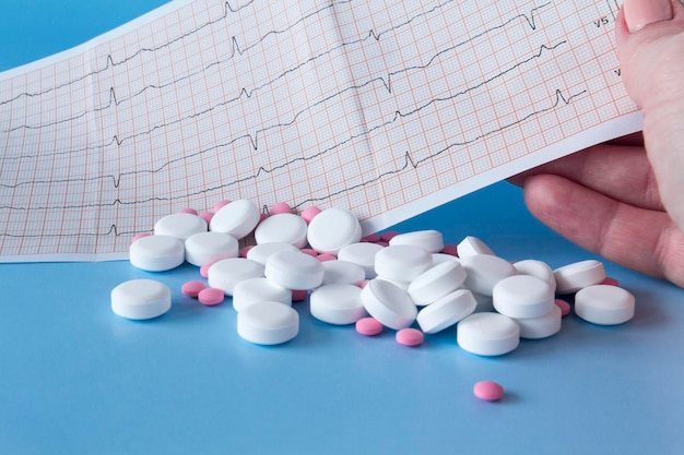 Een groot handvol roze en witte medicijnen op het elektrocardiogram van het hart en een vrouwelijke hand op een blauwe achtergrond Het concept van een gezonde levensstijl en tijdig medisch onderzoek