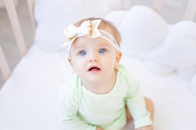 Een groot portret van een schattig klein babymeisje met grote blauwe ogen die in de camera kijken met een strik op haar hoofd op een wieg in een groene katoenen bodysuit