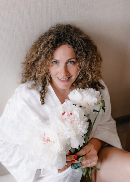 Een groot portret van een vrouw in een witte jurk met krullend haar met een boeket witte pioenrozen in haar hand.