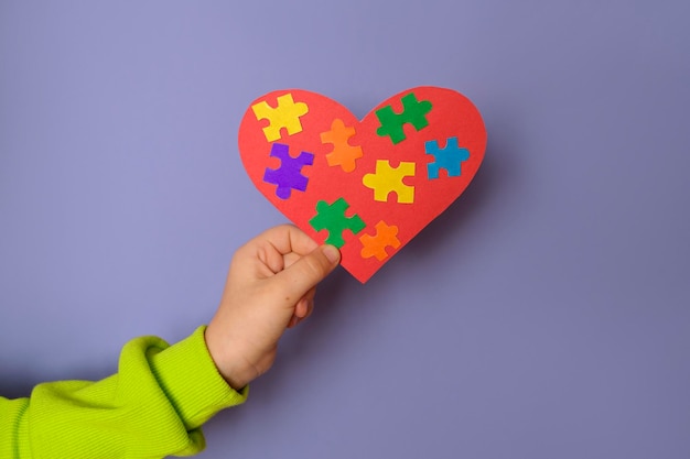 Een groot rood hart met details van gekleurde puzzels erin, in de hand van een kind staat een symbool van autisme